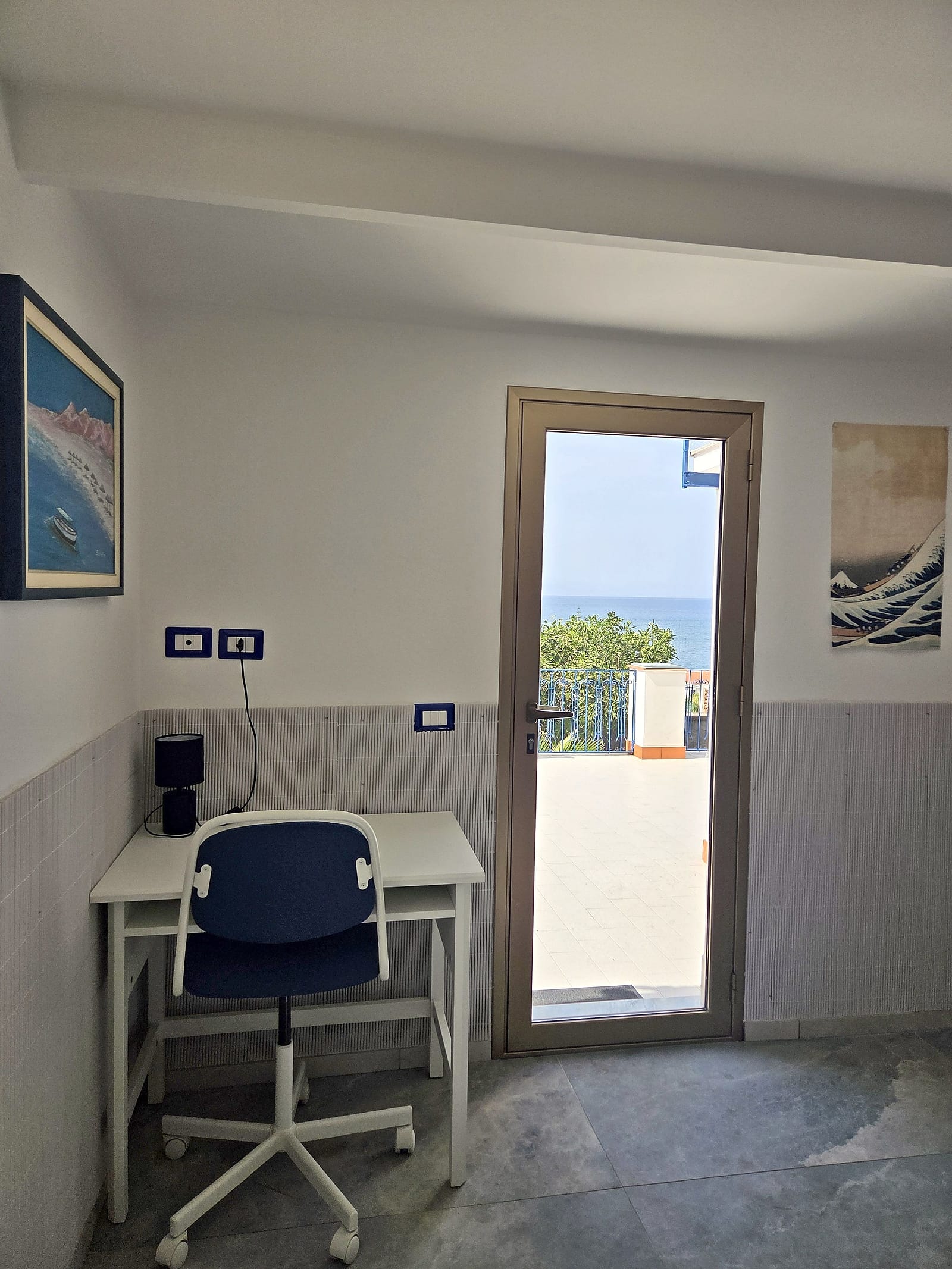 Desk with a blue chair beside the glass door open onto the terrace and the sea