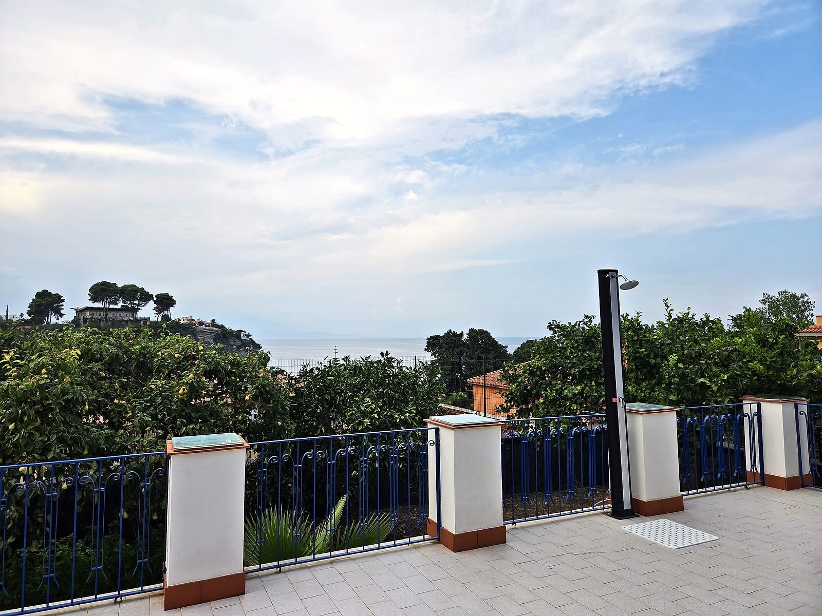The terrace with its outdoor shower, blue railing and a glimpse of the sea beyond the trees